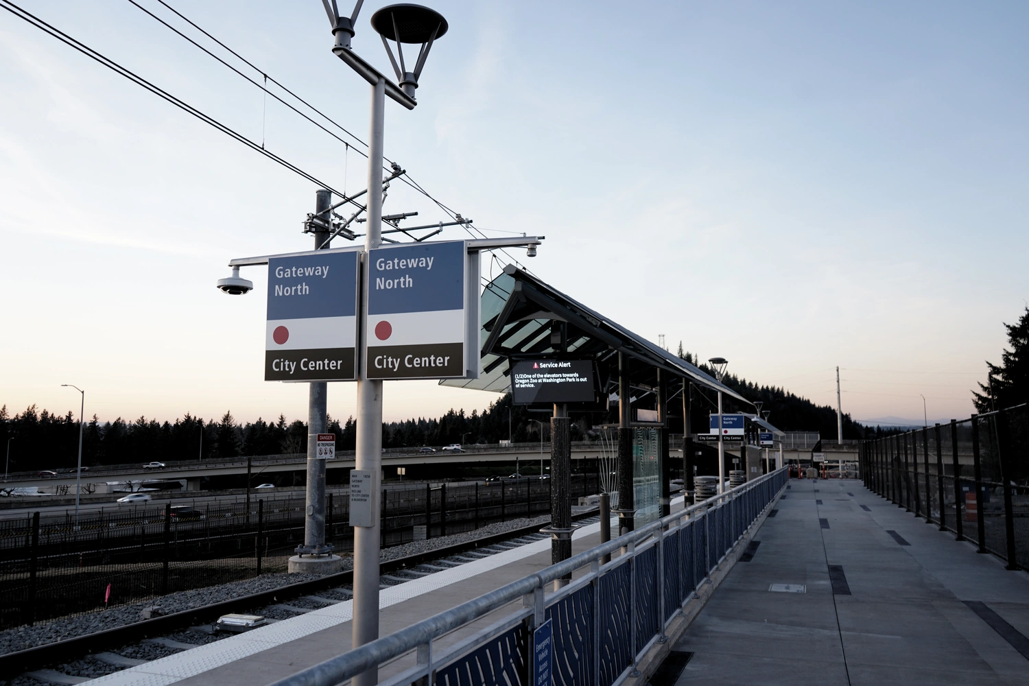 A railway platform shelter, platform, track, and catenary pole lie on
       the left;
       in the foreground is a sign that reads 'Gateway North' and 'City Center'
       with a red circle between them.