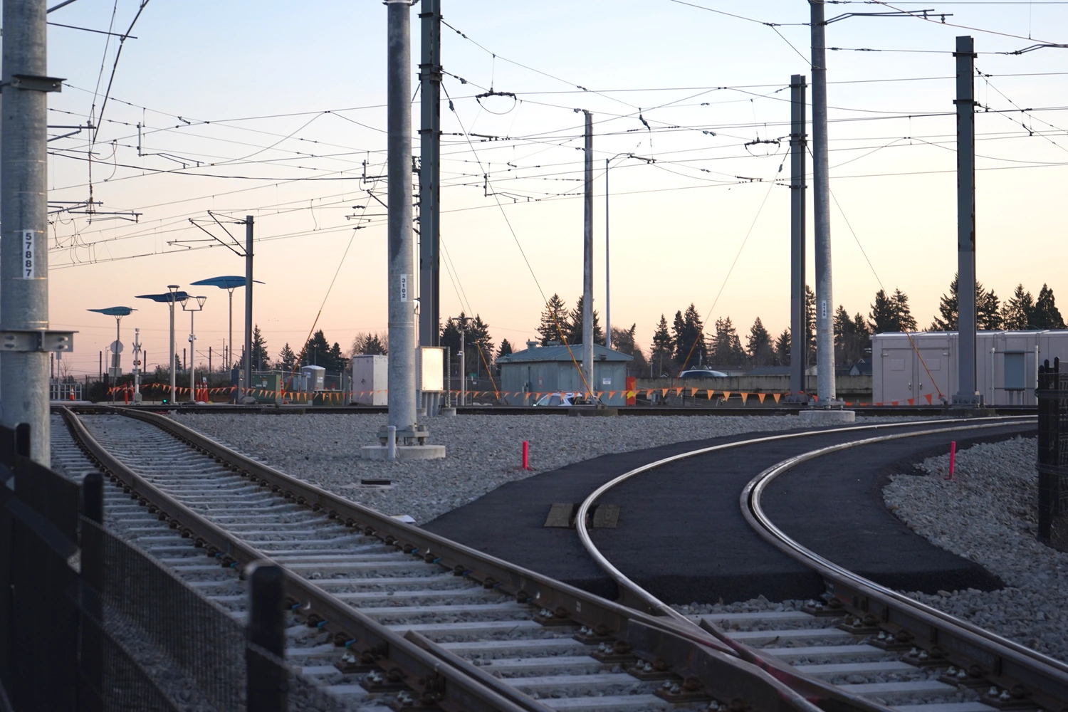 Two tracks with overhead catenary electrification form part of a
       wye junction; one continues straight away
       from the camera, while another curves right.
       The ballast and ties look new.
       Beyond these tracks lie assorted construction materials, the other tracks
       in the junction, buildings,
       and a platform shelter.